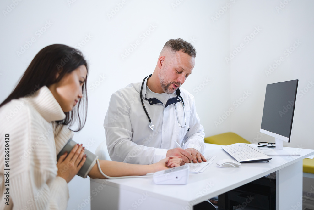 The male doctor measures the woman blood pressure and records the results in the patient's card. Female at the appointment at cardiologist The male doctor measures the woman blood pressure and records the results in the patient's card. Female at the appointment at cardiologist