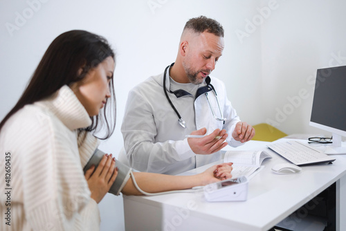 The male doctor measures the woman blood pressure and records the results in the patient's card. Female at the appointment at cardiologist