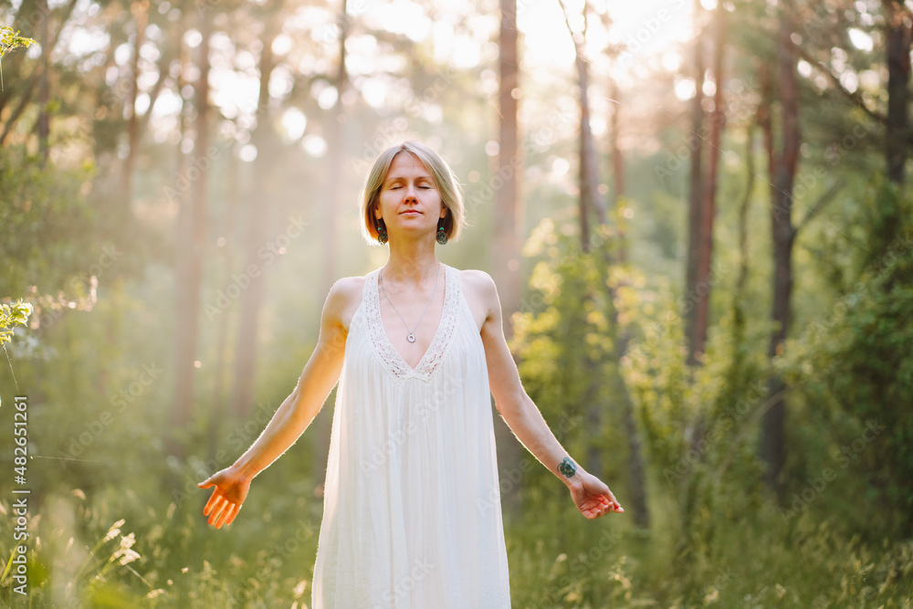 Unity with nature. Young woman in white nightgown standing in green forest on sunny morning with closed eyes and outstretched arms, female breathing deep fresh air in meadow at sunrise. Forest Bathing Unity with nature. Young woman in white nightgown standing in green forest on sunny morning with closed eyes and outstretched arms, female breathing deep fresh air in meadow at sunrise. Forest Bathing