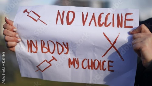 Close-up anti-vax placard in Caucasian female hands with blurred urban city at background. Unrecognizable woman protesting against mandatory Covid-19 vaccination and green pass outdoors photo