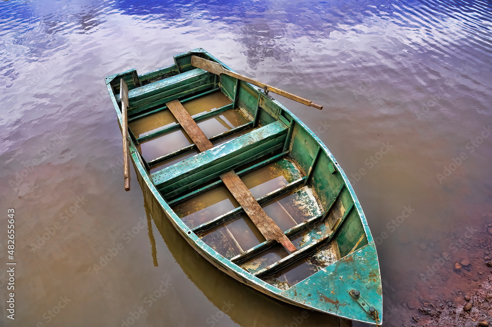 Bote de remos abandonado con agua en su interior. En un río de color marrón que al fondo se hace azul por el reflejo del cielo.