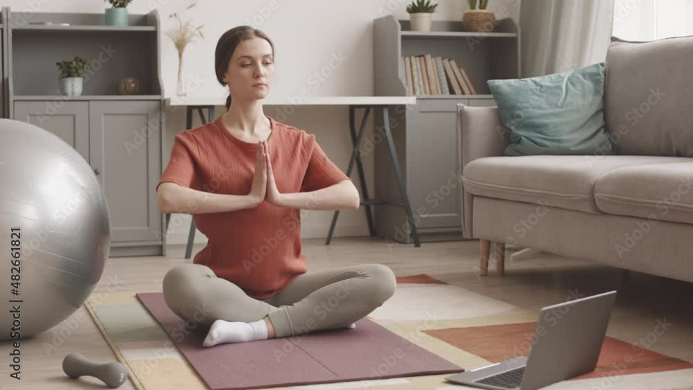 Stab shot of young pregnant woman watching yoga lesson on laptop, sitting on mat in lotus positing with praying hands and her eyes closed Stab shot of young pregnant woman watching yoga lesson on laptop, sitting on mat in lotus positing with praying hands and her eyes closed