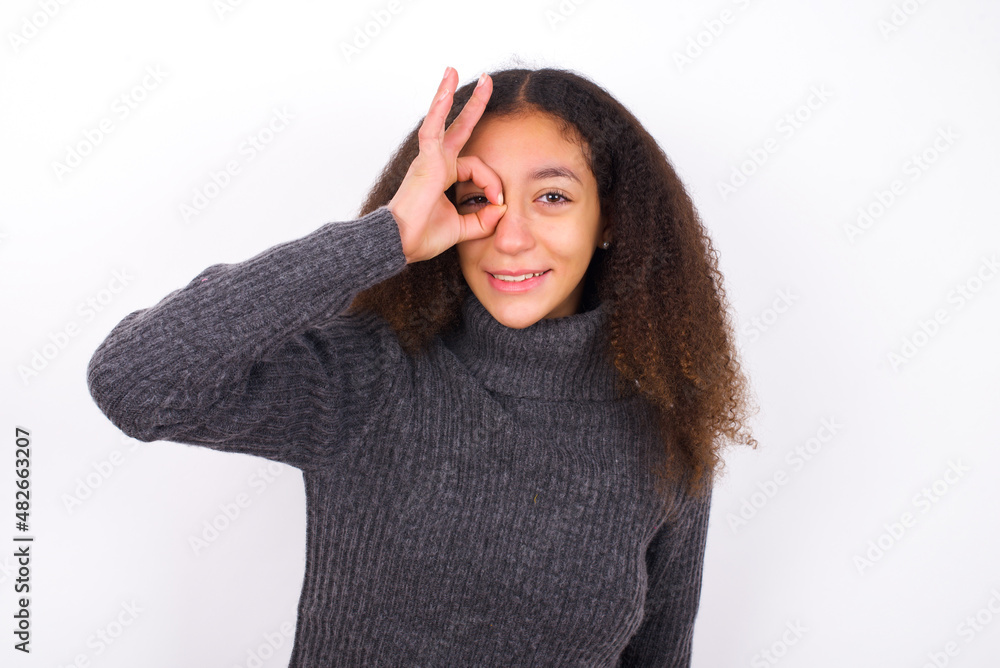 teenager girl wearing grey sweater standing against wite background doing ok gesture with hand smiling, eye looking through fingers with happy face. teenager girl wearing grey sweater standing against wite background doing ok gesture with hand smiling, eye looking through fingers with happy face.