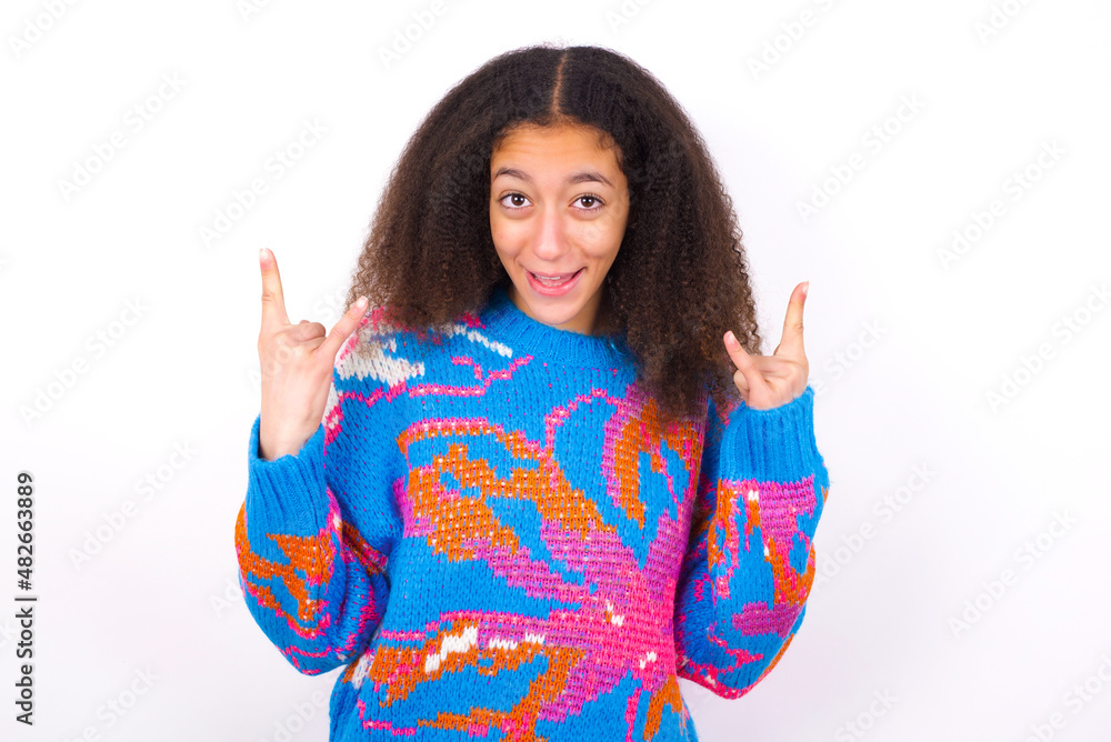 beautiful teenager girl wearing colorful sweater standing against wite background makes rock n roll sign looks self confident and cheerful enjoys cool music at party. Body language concept. beautiful teenager girl wearing colorful sweater standing against wite background makes rock n roll sign looks self confident and cheerful enjoys cool music at party. Body language concept.