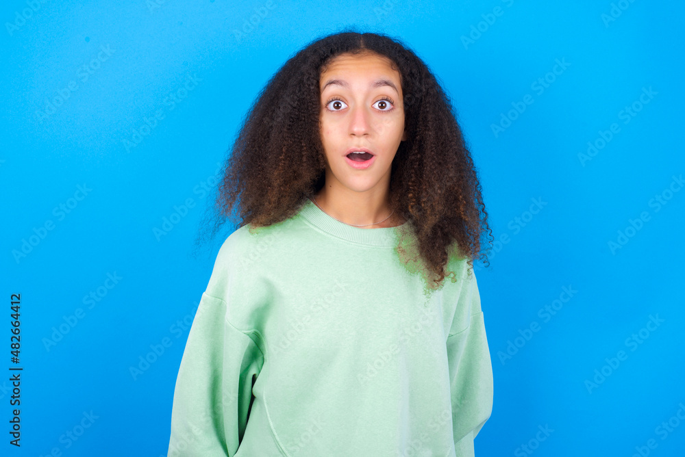 Fototapeta premium Shocked beautiful teenager girl wearing green sweater standing against blue background stares bugged eyes keeps mouth opened has surprised expression. Omg concept