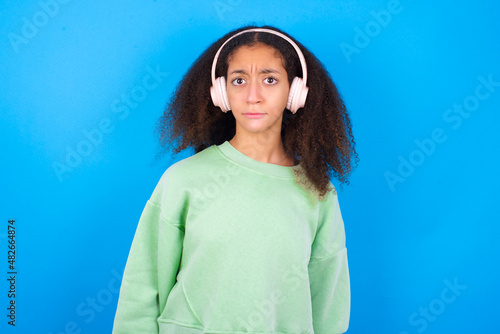 Serious displeased beautiful teenager girl wearing green sweater standing against blue background looks puzzled at camera being angry wears stereo headphones listens music while walking at street