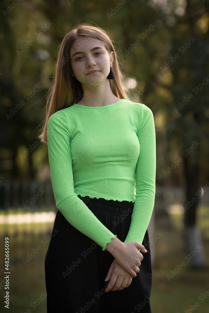 Portrait of a young beautiful blonde girl in a yellow-green blouse and black pants in the park.
