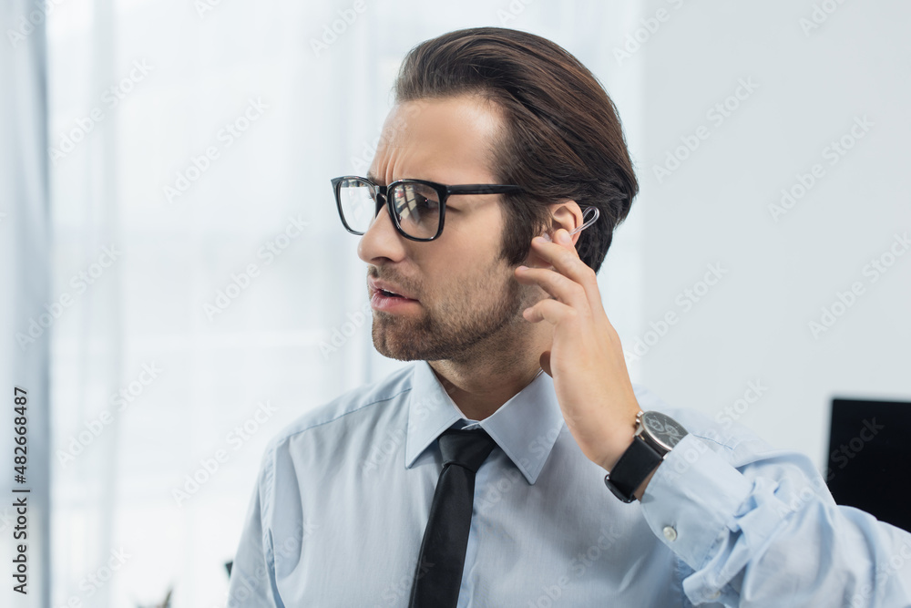 serious security man in eyeglasses touching earphone while working in office.
