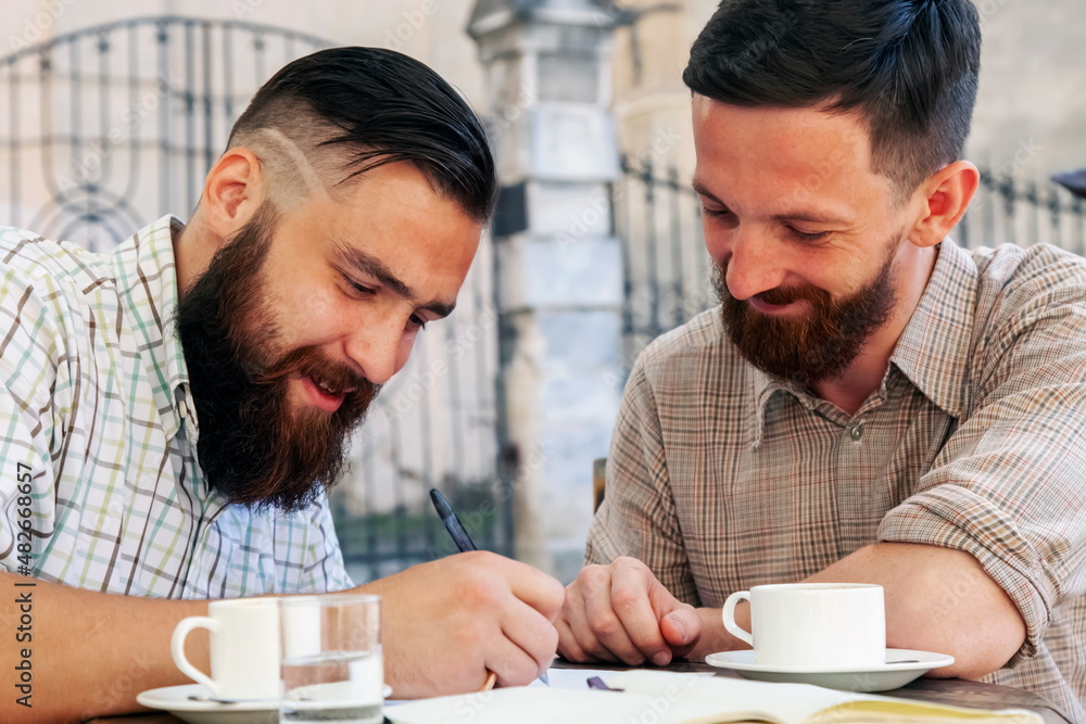 Two hipster freelance businessmen working outdoors on the project and drinking coffee. Concept: happiness, freedom and favorite job. Horizontal image. Two hipster freelance businessmen working outdoors on the project and drinking coffee. Concept: happiness, freedom and favorite job. Horizontal image.