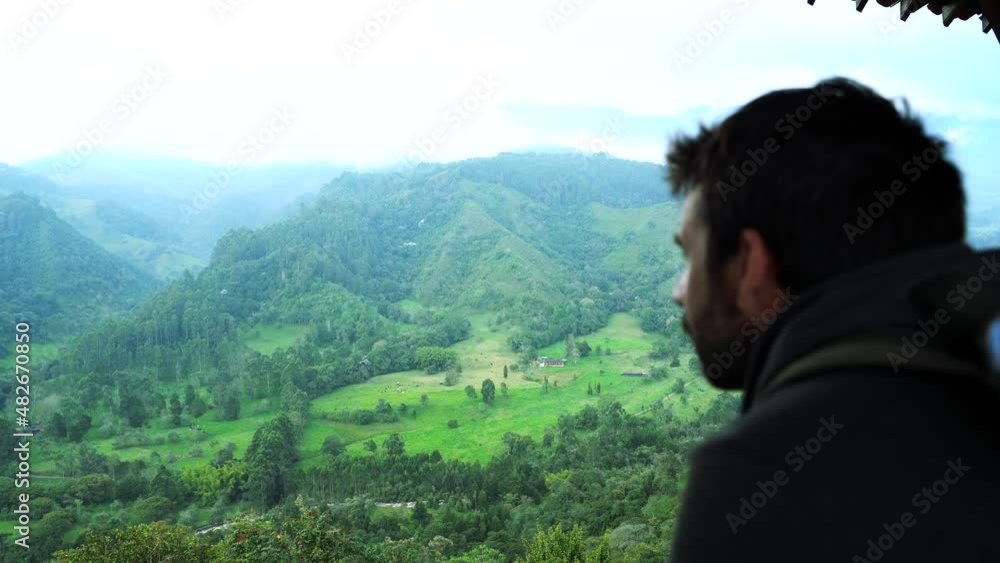 Rear view of unrecognizable man sightseeing on porch at valley in Colombia. Horizontal panoramic view of man with backpack traveling in Filandia. People and travel destination in South America. Rear view of unrecognizable man sightseeing on porch at valley in Colombia. Horizontal panoramic view of man with backpack traveling in Filandia. People and travel destination in South America.