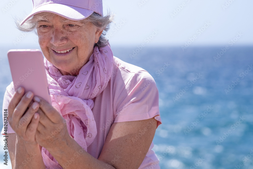 Senior adult caucasian attractive woman pink dressed sitting close to the sea in a sunny day, using mobile phone . Relaxed pensioner female enjoying outdoor and good time using wireless technology