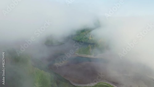 Flying through white clouds over dark sea with green hills at deserted shoreline
