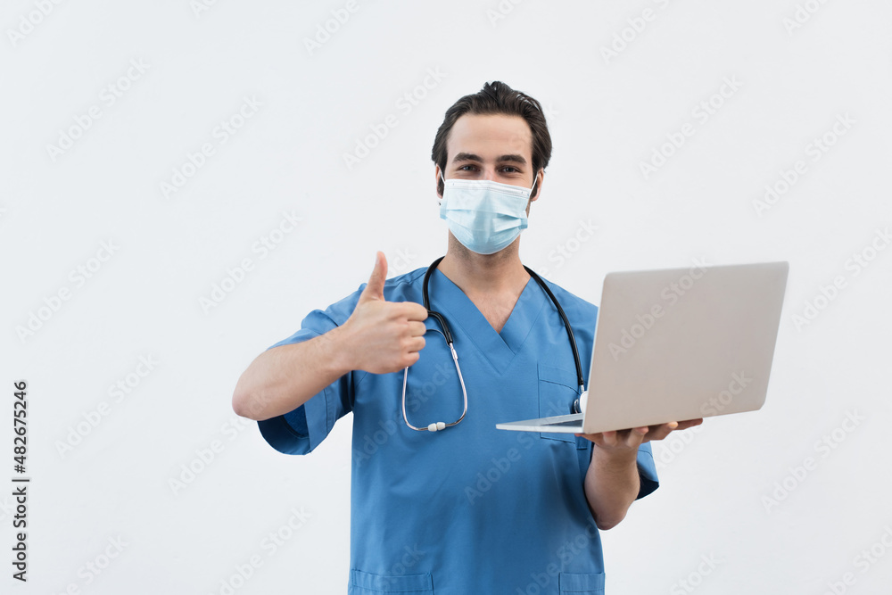 young doctor in medical mask holding laptop and showing like gesture isolated on grey. young doctor in medical mask holding laptop and showing like gesture isolated on grey.