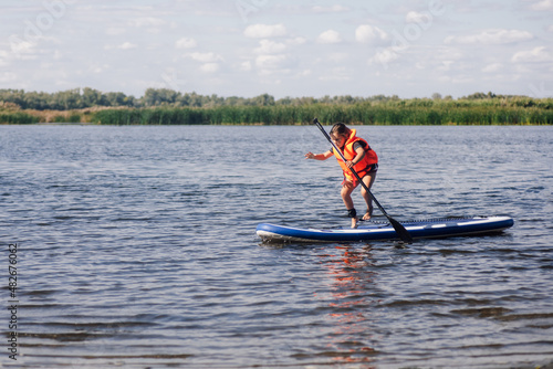 Little female child standing on paddle board on clue lake trying to keep balance on feet wearing orange life jacket. Active holidays full of adventures. Inculcation of love for sports from childhood.