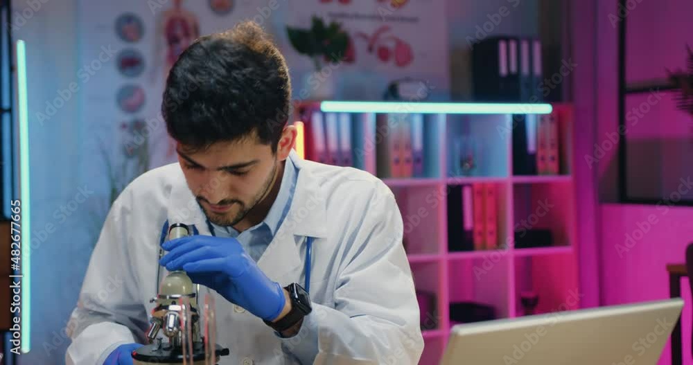 Likable confident qualified young bearded researcher in uniform working with microscope and computer during overtime work in evening laboratory Likable confident qualified young bearded researcher in uniform working with microscope and computer during overtime work in evening laboratory
