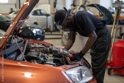 Young African man in workwear bending over open car hood while checking engine or repairing details of vehicle
