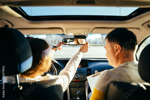 Close up of female hands correcting mirror. Traveler girl on car trip, looking at the road. View over shoulder Female on test drive car with instructor photo