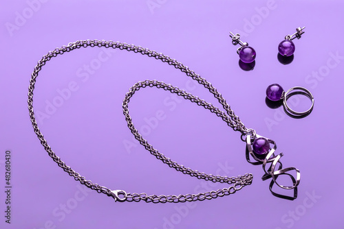 Bijouterie on a violet glossy background. Imitation jewelry. Earrings, chain pendant, ring with purple round stone. Women's glamour accessories. Fashion concept. photo