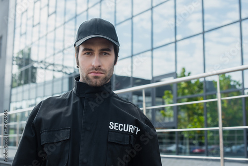 young security man in black uniform looking at camera near building with glass facade.