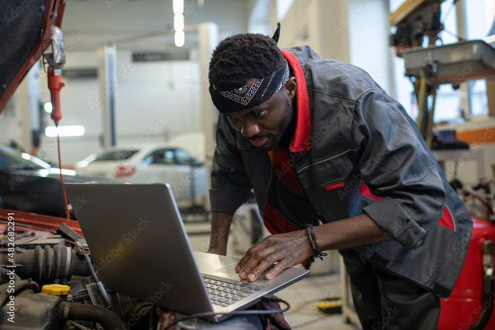 Young African repairman in workwear consulting websites while checking and repairing engine of car