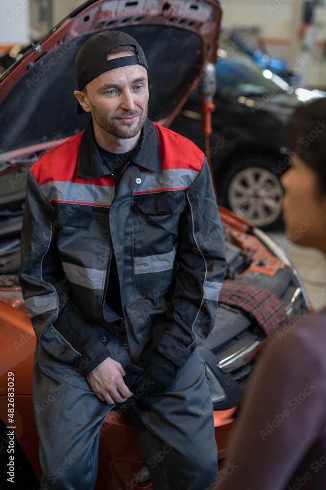 Young contemporary automechanic in workwear listening to female colleague while standing against open hood of car