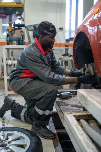 Young professional automechanic of African ethnicity in workwear and protective gloves changing wheel of car