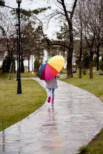 A cute little girl in a blue cape  pink boots and a pink hat runs through puddles and has a fun.The girl has a rainbow umbrella in her hands. Happy childhood. Early spring. Emotions.
