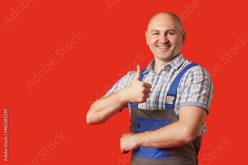 Smiling, happy, bald, unshaven man in work clothes shows an ok hand gesture on a red background. The worker has completed the work.