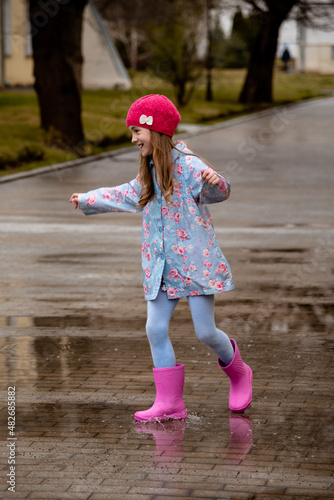 A cute little girl in a blue cape, pink boots and a pink hat runs through puddles and has a fun. Happy childhood. Early spring. Emotions.