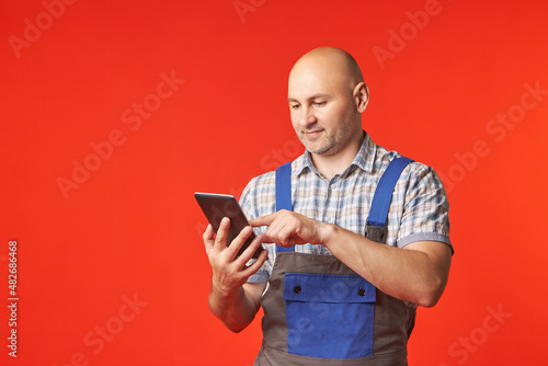 Bald man in a work suit holds a phone with one hand and works in it with the other, a photo on a red background. The employee accepts orders using the phone