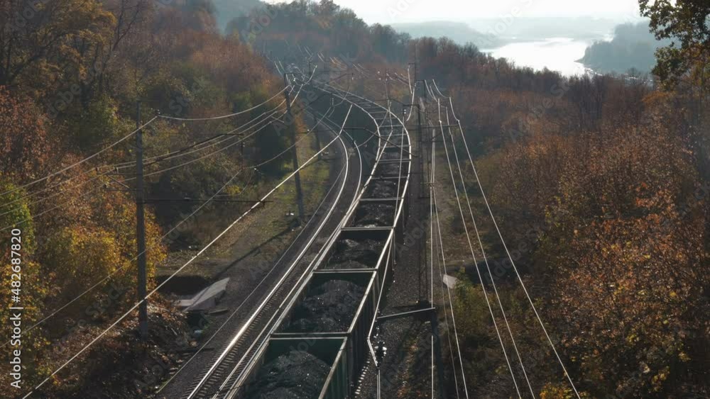 Freight train carries coal an electric locomotive by two-sided railway along the river and colorfull forest at sunny autumn day. Aerial top view Freight train carries coal an electric locomotive by two-sided railway along the river and colorfull forest at sunny autumn day. Aerial top view