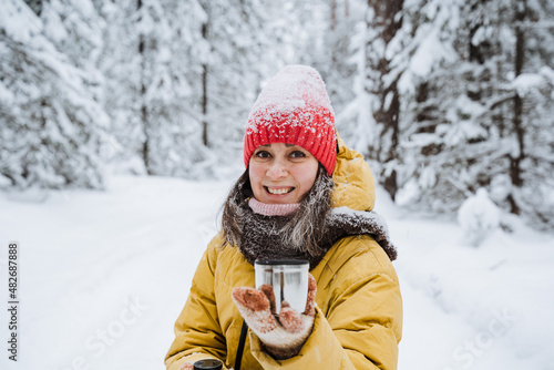 A young girl smiles in the winter forest, a mug of coffee in her hand. The red cap is covered with snowflakes, the weather is cold. A woman in a yellow jacket walks through a snowy forest.