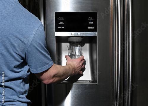 Male right hand filling glass with water splashing out of dispenser of home fridge photo