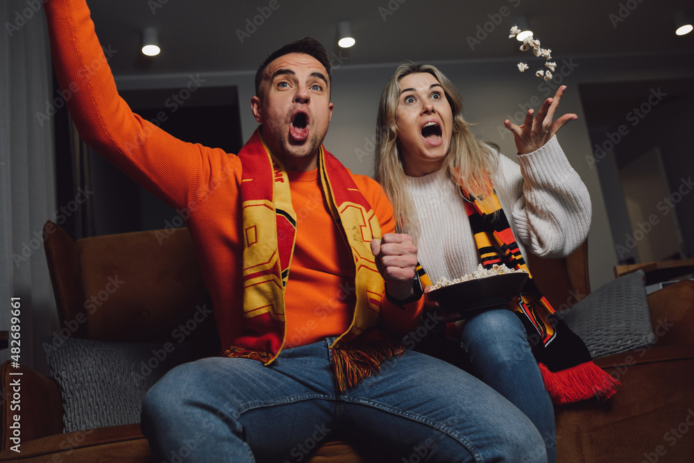 Dark-haired smiling enthusiastic man and blonde woman in scarf watching socker victory goal on TV on comfort couch.