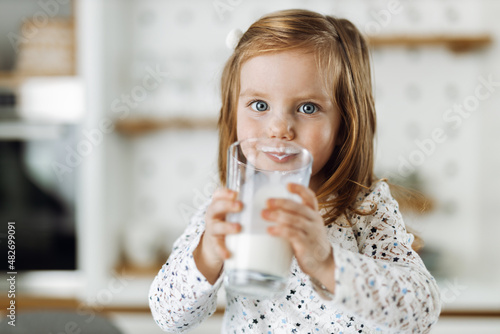 Foto Cute little girl drinking fresh milk at home and looking at camera