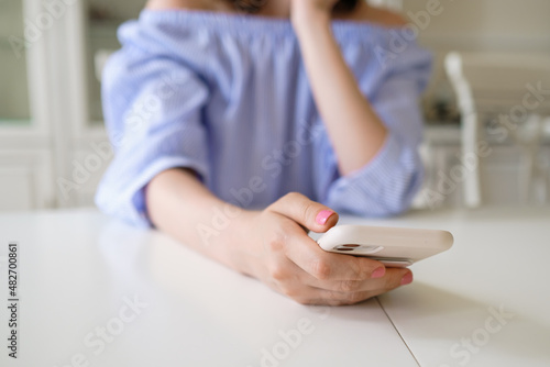 Young woman in stylish blue blouse with neat manicure holds modern smartphone sitting at white table in morning in flat room closeup