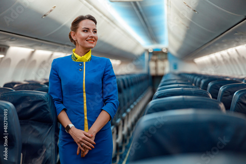 Cheerful woman stewardess in airline air hostess uniform standing in aisle of airplane passenger salon photo