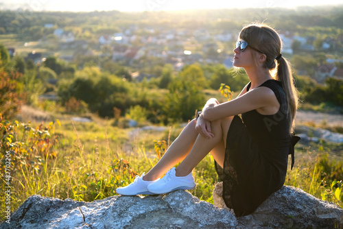 Young elegant woman in black short dress sitting on a rock relaxing outdoors at summer evening. Fashionable female enjoying warm sunset in nature