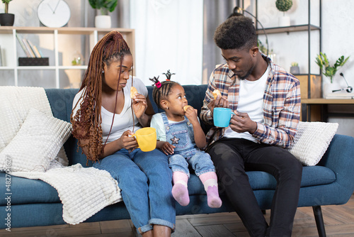 African americna parents and little daughte rdrinking hot tea and eating fresh croissants. Happy family of three enjoying snack break on comfy couch at home. photo
