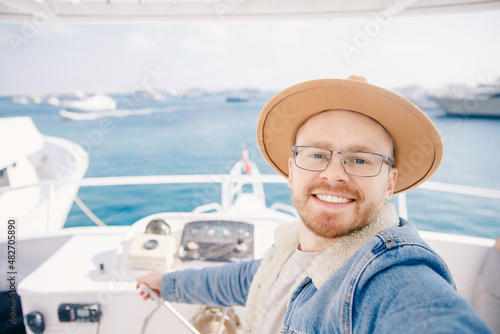 Happy young man in hat goes on trip on white yacht sea making selfie photo on camera, concept travel life hipster
