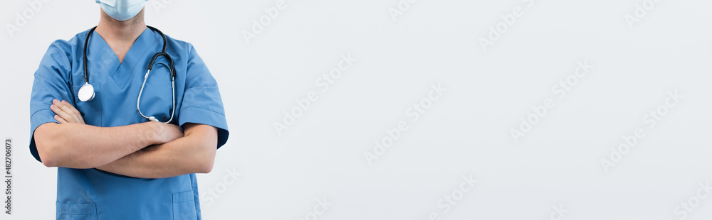 partial view of physician in blue uniform and medical mask standing with crossed arms isolated on grey, banner. partial view of physician in blue uniform and medical mask standing with crossed arms isolated on grey, banner.