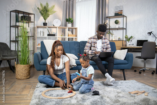African american man sitting on couch and typing on laptop. Young woman using various toys for playing with daughter on floor. Domestic lifestyles of happy family.