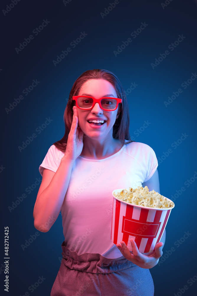 Smiling young woman wearing 3d glasses holding popcorn bucket standing on dark blue studio background.