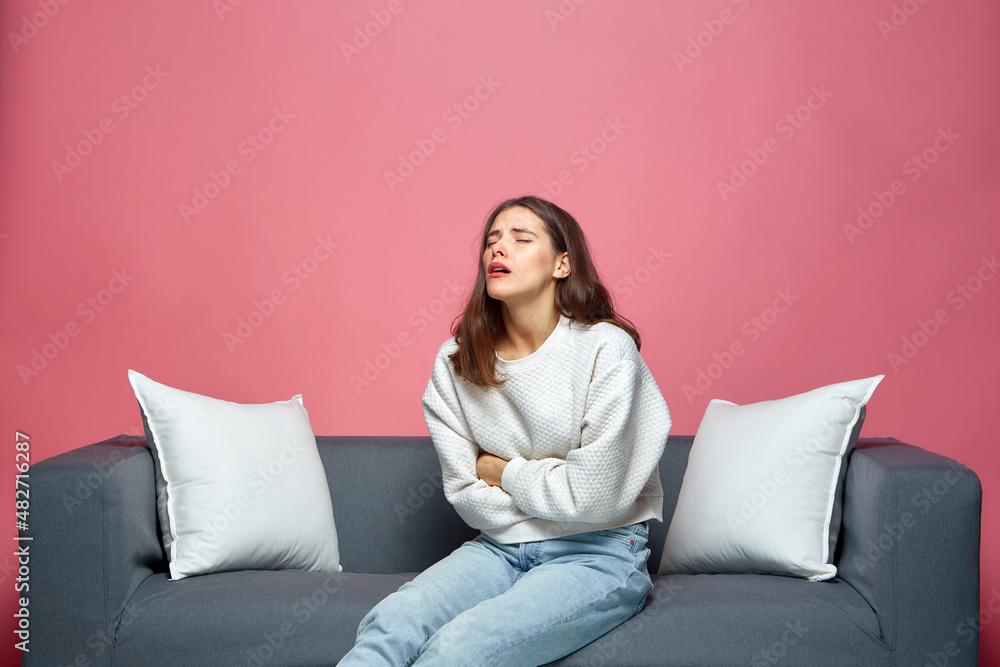 Unhealthy girl touch stomach, suffering abdominal pain, sitting on sofa. Nausea, stomach ache, painful periods Unhealthy girl touch stomach, suffering abdominal pain, sitting on sofa. Nausea, stomach ache, painful periods