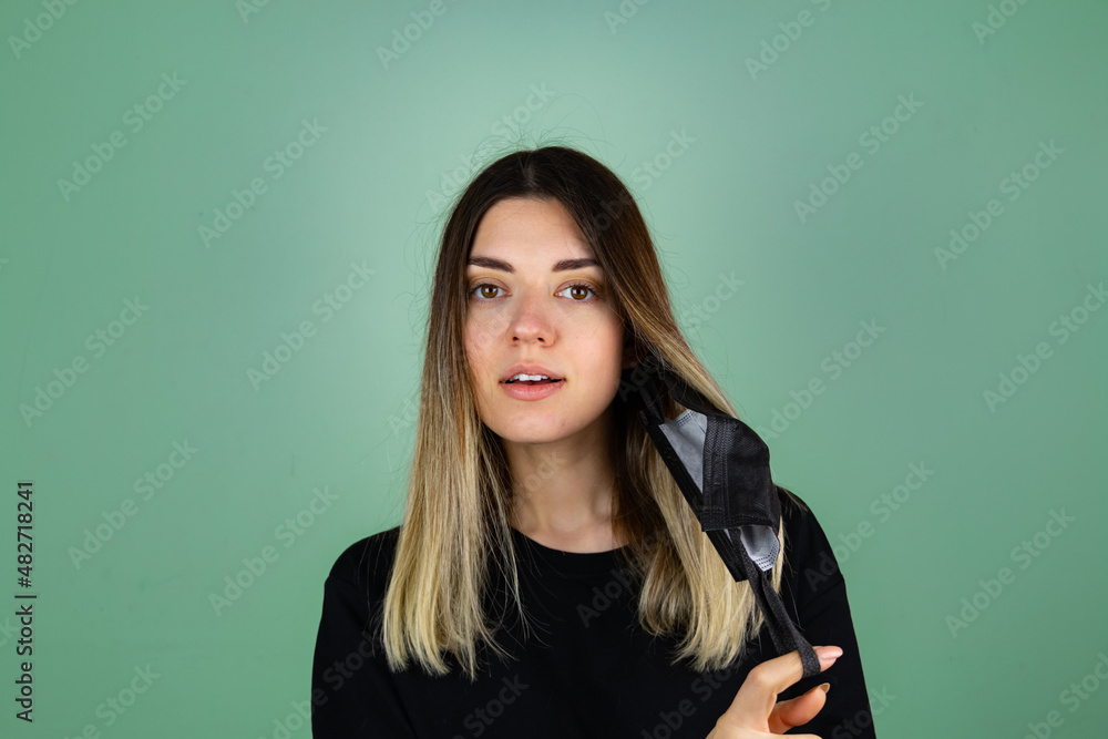 Young woman takes off protective mask and looking at camera while corona virus pandemic. Quarantine, and social distance concept. Green isolated studio background. Young woman takes off protective mask and looking at camera while corona virus pandemic. Quarantine, and social distance concept. Green isolated studio background.
