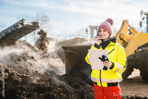 Manager in biomass and landfill operation using her radio in front of machines, clipboard in hand