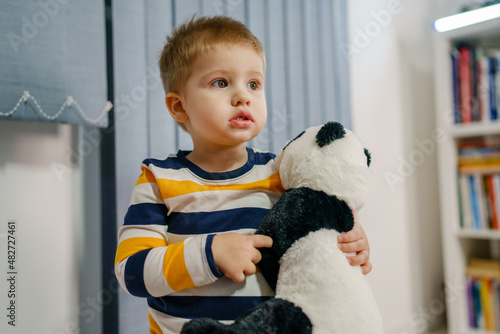 Waist up portrait of one small caucasian boy two years old holding panda toy while standing alone at home looking to the side copy space childhood and growing up concept photo