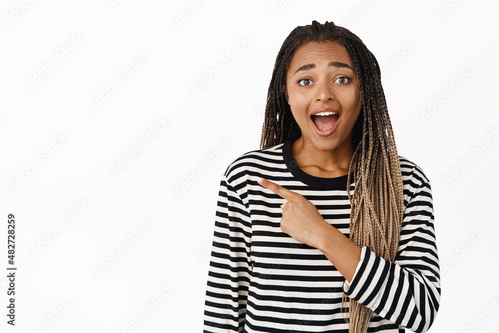 Happy black girl laughs over funny advertisement, points finger left, shows promo sale, stands over white background, isolated shot Happy black girl laughs over funny advertisement, points finger left, shows promo sale, stands over white background, isolated shot