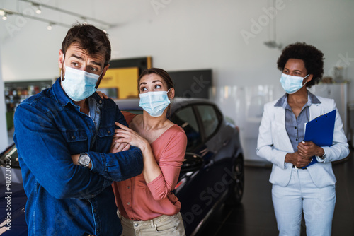 Young couple with protective face masks on their faces buying new car at car showroom.