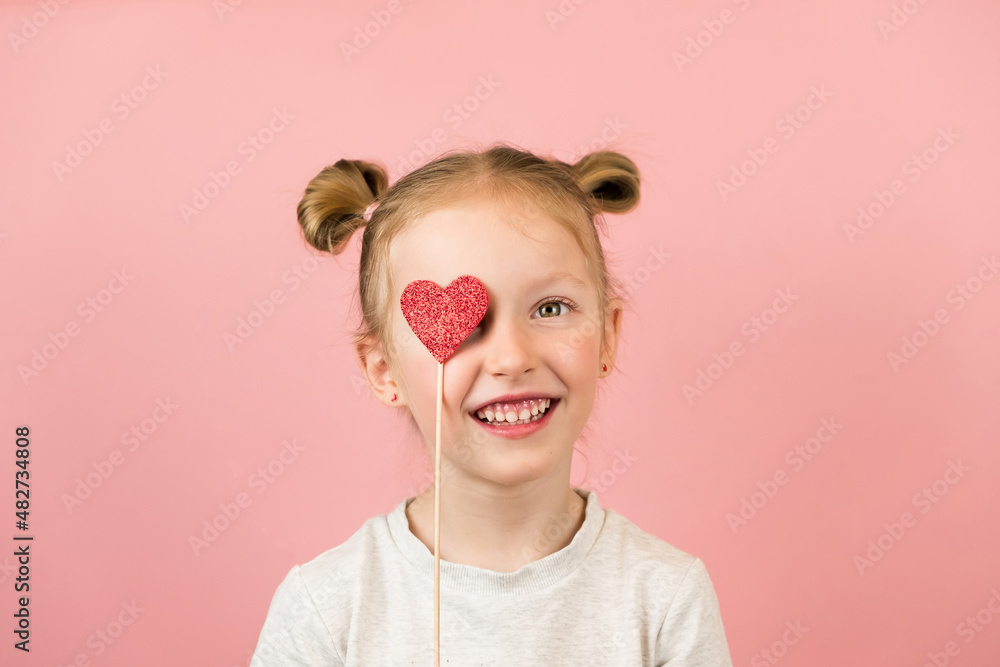 Funny little blonde girl smiling and playing with red heart toy on pink background. Valentines Day or Mothers Day concept Funny little blonde girl smiling and playing with red heart toy on pink background. Valentines Day or Mothers Day concept